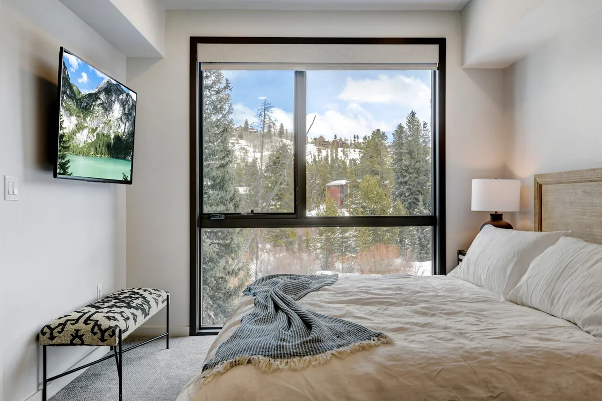 A serene bedroom scene featuring a large window with mountain views, a bed with cozy bedding, a wall-mounted television, and a decorative bench. Natural light fills the space, and the overall atmosphere feels peaceful and inviting.