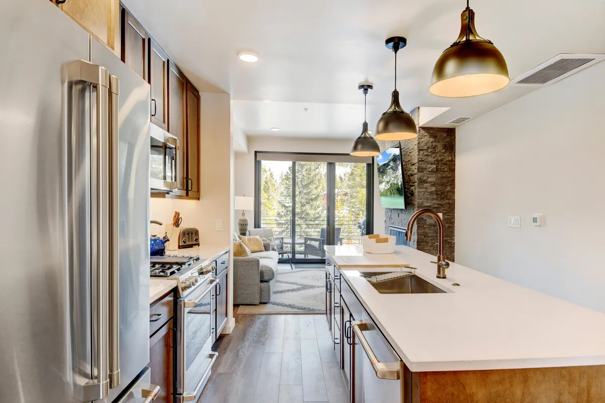 A modern kitchen with expansive windows providing plenty of natural light, featuring an island with a sink, modern kitchen appliances, and views that connect the kitchen to the outdoors. The space is arranged for both cooking and entertaining.