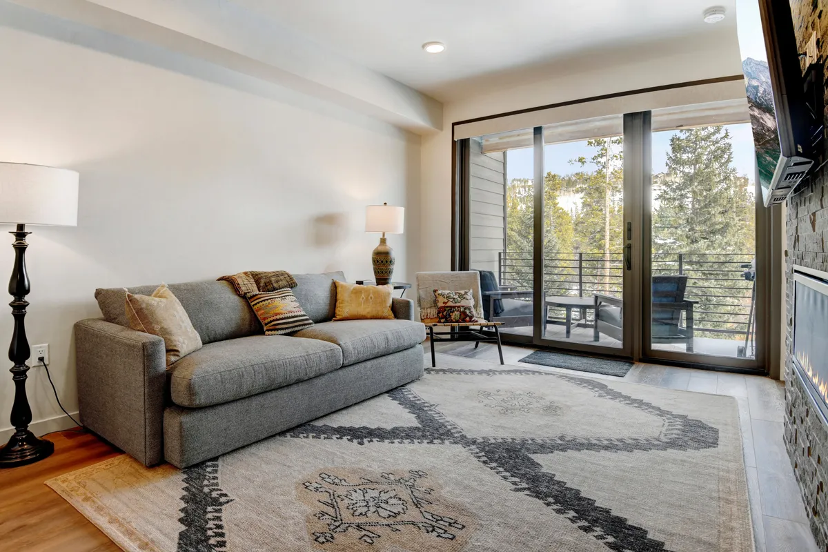 A stylish, serene living space featuring a cozy sofa, contemporary rug, and decorative chair. Sliding glass doors open to a balcony, offering sunset views and inviting natural light. The calm ambiance sets the stage for relaxing, reading, or socializing with friends and family.