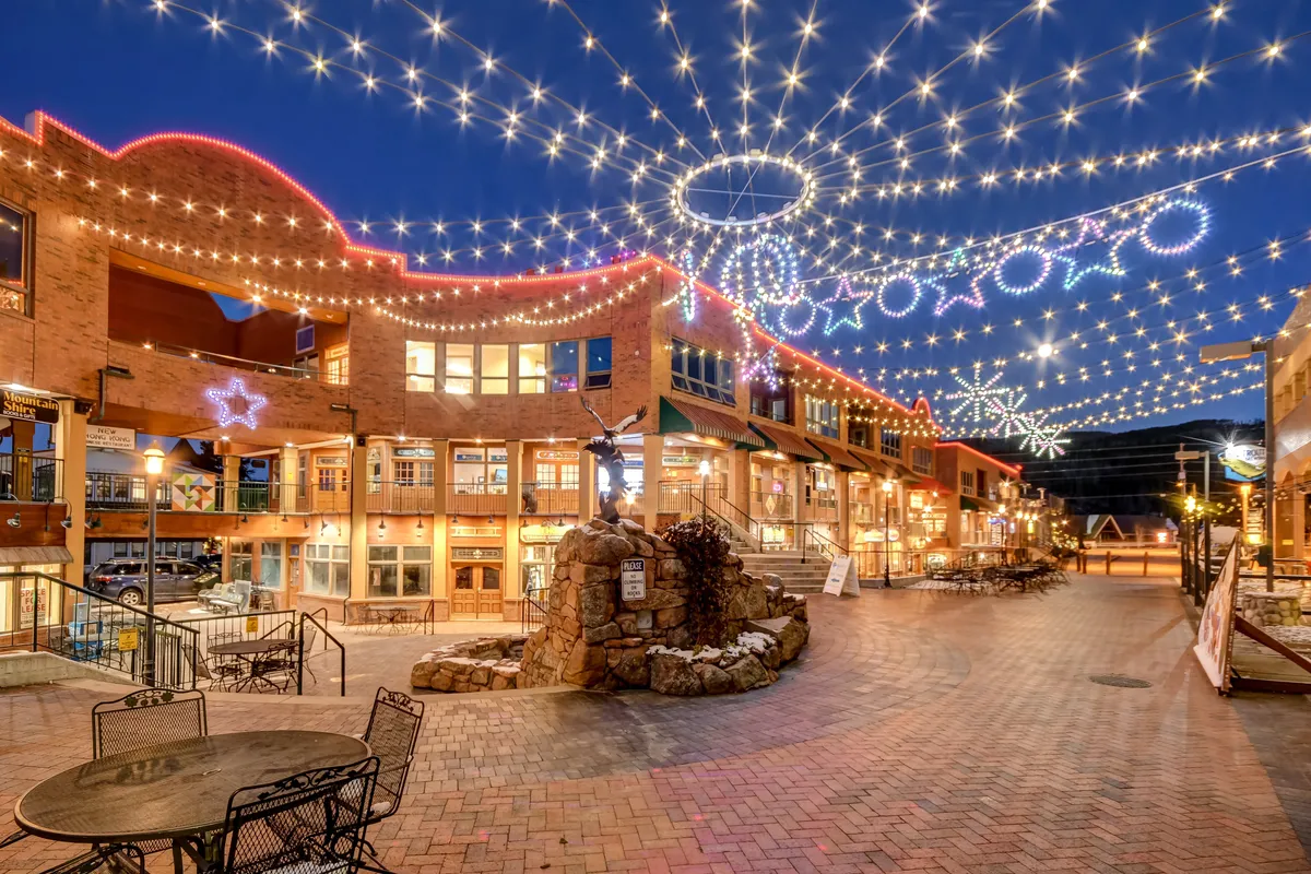 A lively outdoor plaza scene adorned with string lights, decorative stars, a stone fountain, and outdoor seating. People are shown dining, shopping, and socializing, creating a festive and vibrant atmosphere.