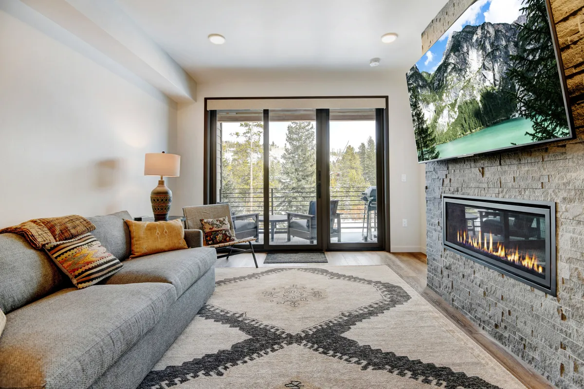 A warm, welcoming living room featuring a plush sofa positioned by a modern fireplace and a large wall-mounted television. Expansive windows flood the space with natural light and frame beautiful mountain scenery, while the carefully selected decor blends contemporary lines with cozy alpine touches.