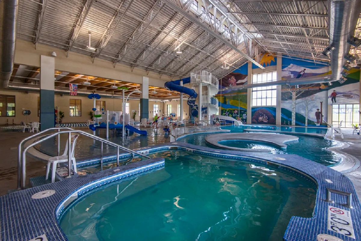The image shows a vibrant indoor recreation area with water slides snaking above large pool areas. A colorful mural decorates the wall, and there are families and children engaged in swimming and water play, suggesting a fun, lively atmosphere suitable for all ages.