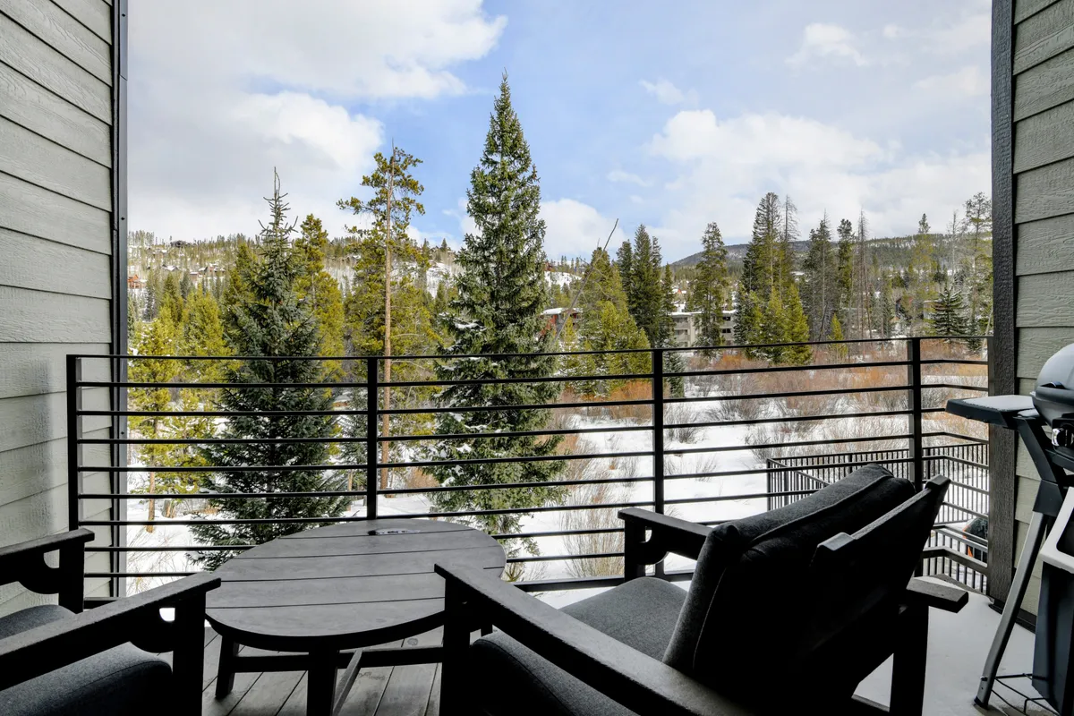 This image captures a tranquil balcony setting overlooking distant mountains and pine trees with snow covering the ground. The balcony is furnished, inviting guests to relax and enjoy the mountain scenery, whether with a morning coffee or a book.