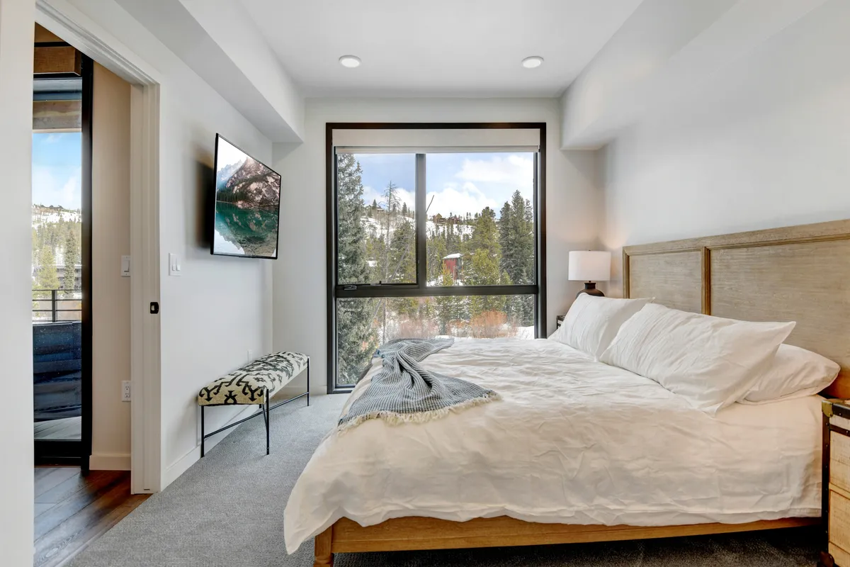 A bedroom with white linens, a large window offering a mountain view, a TV mounted on the wall, and a decorative bench at the foot of the bed. The space feels airy, bright, and restful.