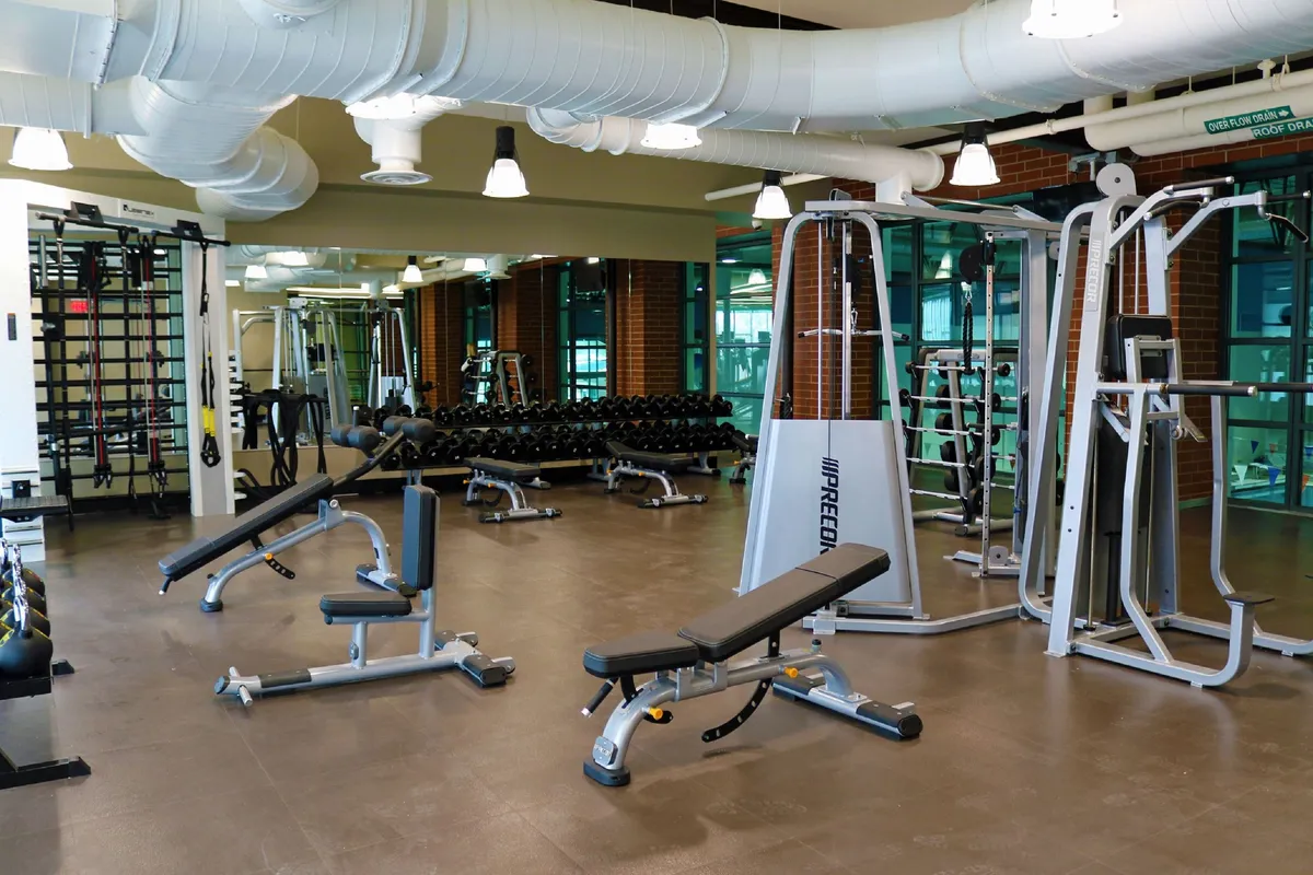 The image displays a modern, fully equipped gym featuring weight benches, dumbbell racks, and exercise machines. The space is well-lit and organized, with a focus on fitness and personal training activities.
