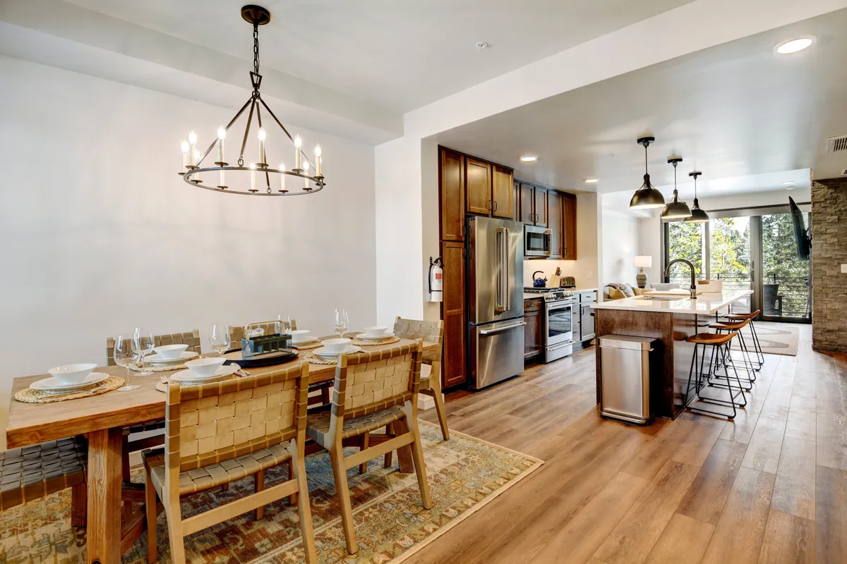 A spacious dining area comes to life with a set dining table under a striking chandelier. The kitchen island—equipped for socializing and casual meals—sits in the background, all illuminated by large windows that reveal mountain scenery. The decor is modern but warm, echoing the section's blend of elegance and alpine charm.