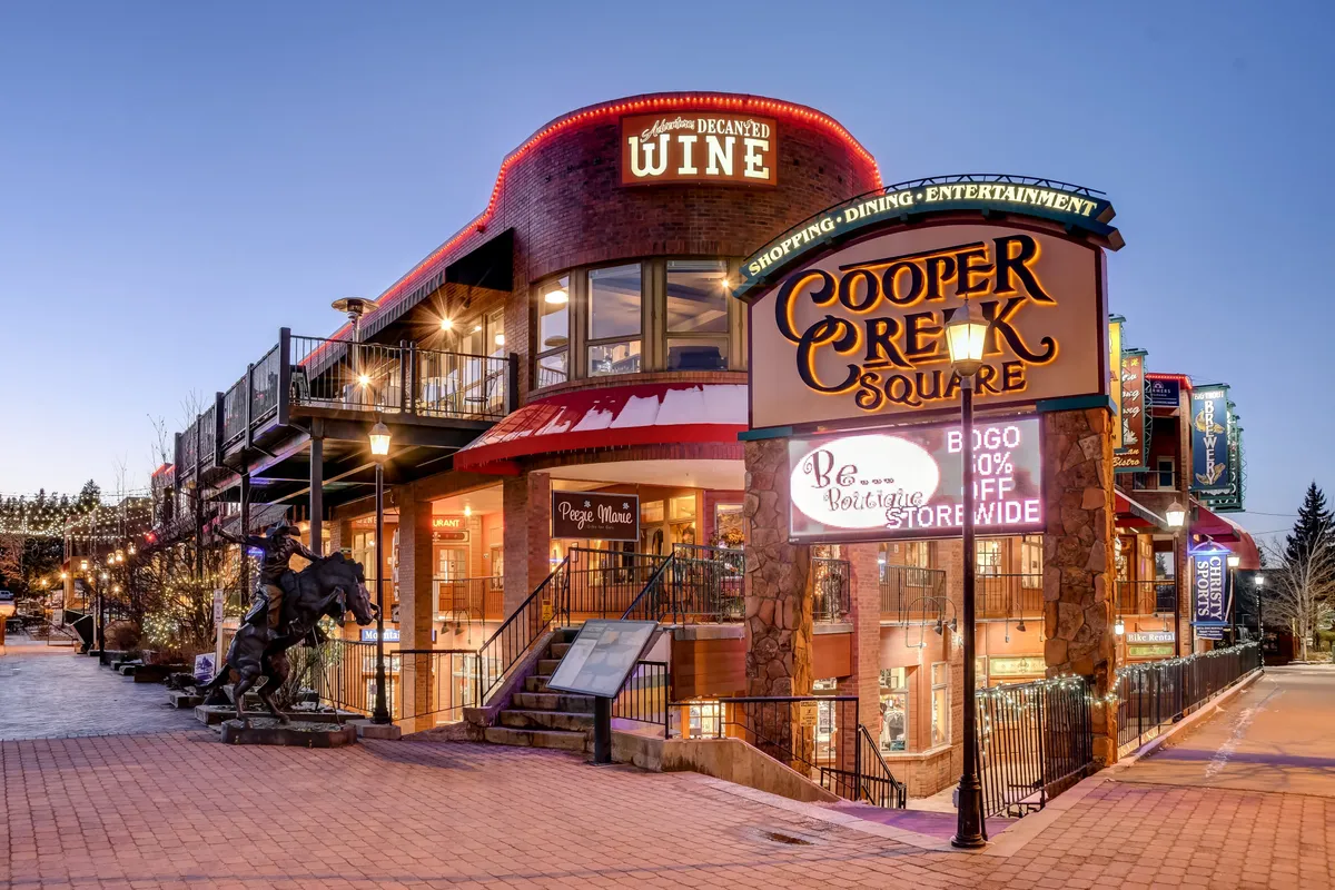 The lively exterior scene at Cooper Creek Square with welcoming signage, wine shop, inviting outdoor seating, and a foreground sculpture. The area exudes a festive, social mood, perfect for families to explore dining, music events, and entertainment.