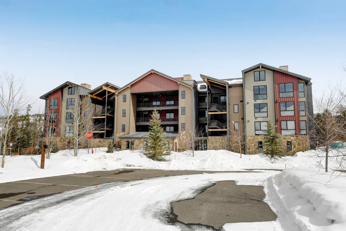 A snowy exterior shot of a modern condo building with warm wooden accents, surrounded by snow-covered trees and a cozy, inviting atmosphere. The scene suggests a peaceful winter retreat, perfect for romantic escapes or unwinding after a day outdoors in the Rockies.