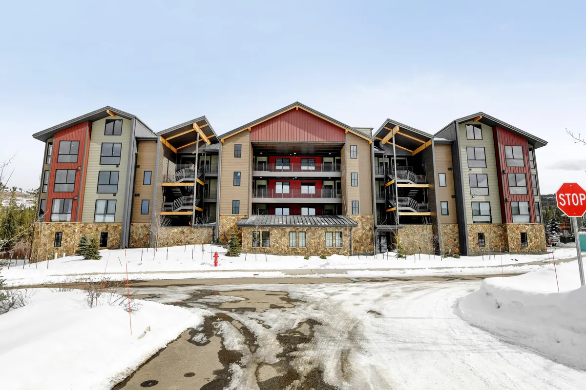 The exterior of the building with its welcoming facade, balconies, and a snowy ground, set against stunning mountain views. People can be imagined gathering or relaxing on the balconies, hinting at the sense of community and year-round outdoor activities.