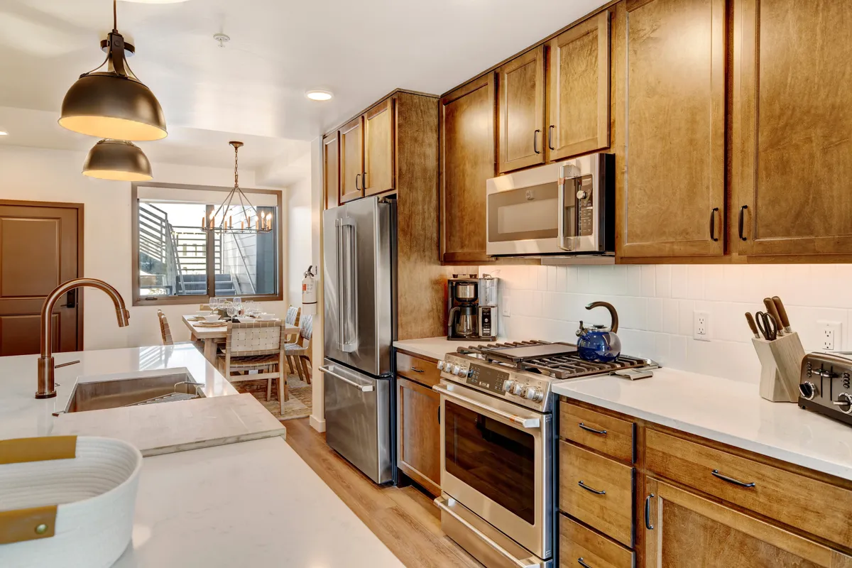 A warmly lit kitchen highlighting modern appliances, wood cabinetry, marble countertops, and a partial view of the adjacent dining area. The space feels open, functional, and ideal for group meal preparations or casual family breakfasts.
